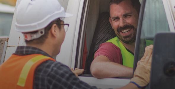 A truck driver leans out his window and talks to a man in a hard hat.