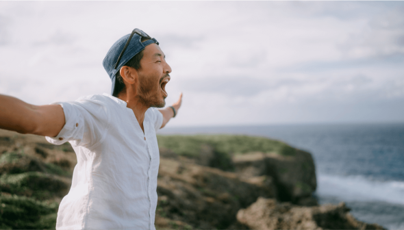 A man looking out at the sea, over a cliff, with arms stretched wide, expressing freedom.