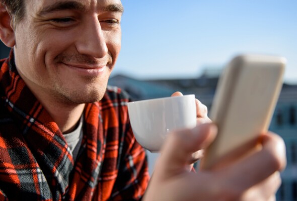 man wearing red and black checkered shirt smiling at his mobile holding a cup in his hand