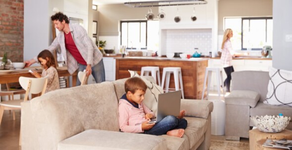 a family of four in their home with boy on a laptop, mum in the kitchen and daughter and dad on the dining table