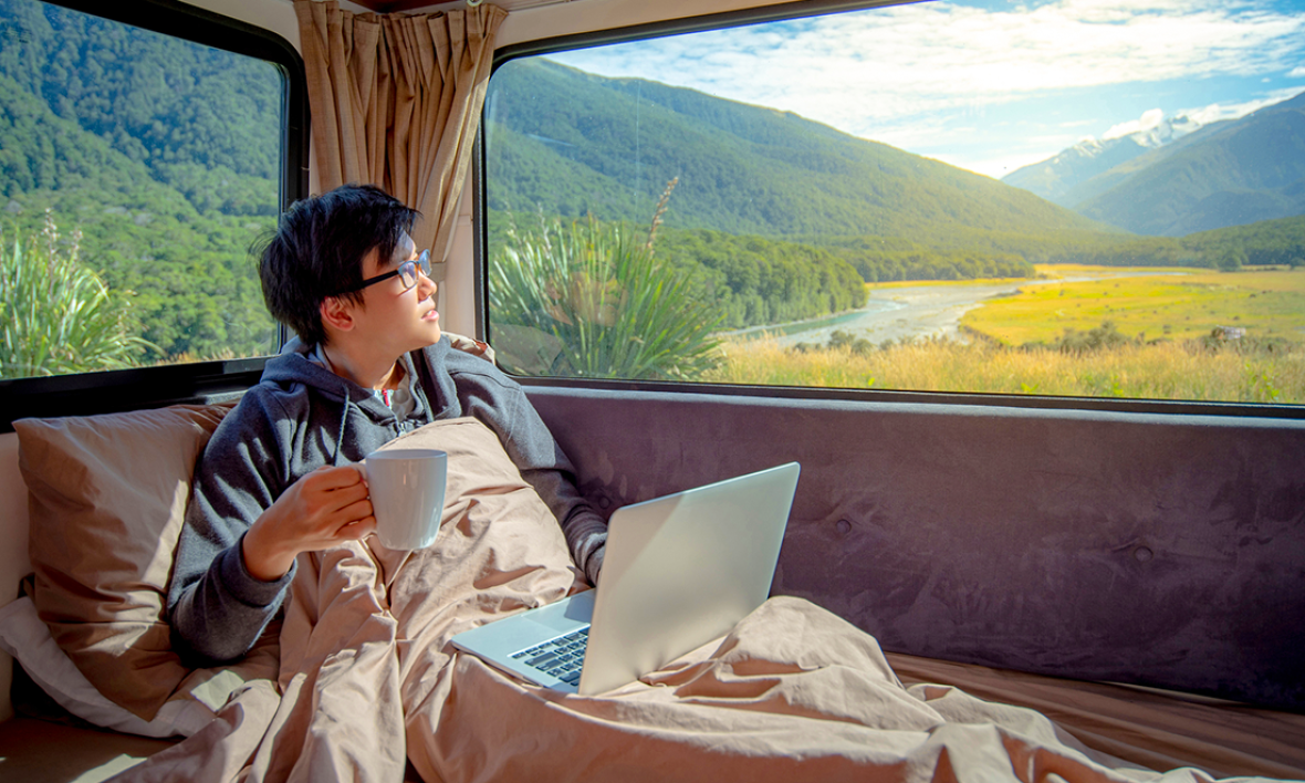 A man with his laptop, looking out the window of his caravan, surrounded by NZ wilderness.