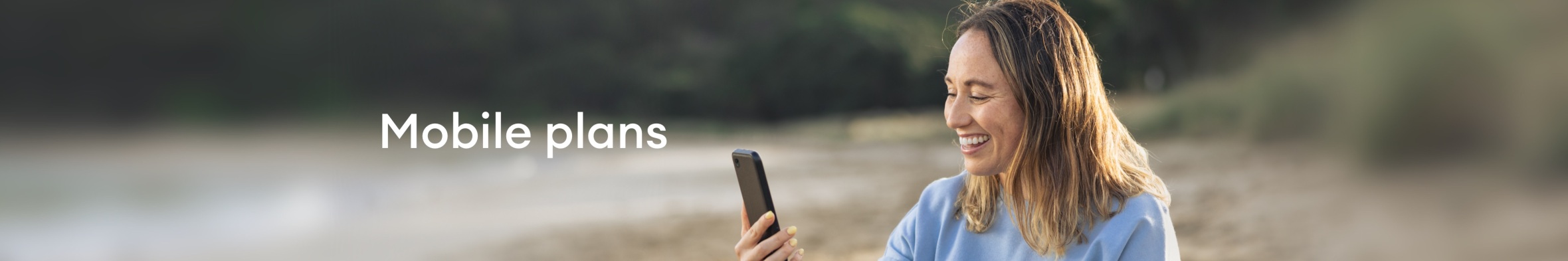 A lady smiling at what's showing on her mobile phone, as she sits on a quiet beach.