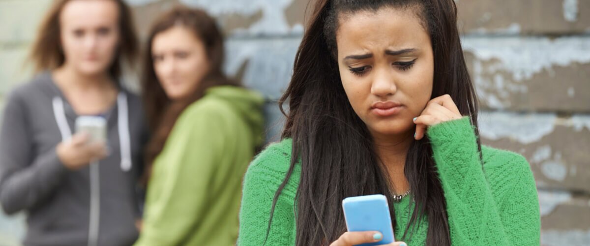 A worried teenager in a schoolyard looking at her phone, with two girls in the background.