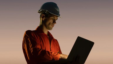 Man outside, working on a laptop, wearing a hardhat.