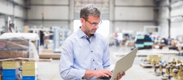 A man stands in the middle of a warehouse looking at his laptop screen while pretending to type. His fake typing fools nobody. Hes killing time til lunchbreak.