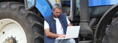 Guy in blue vest next to a tractor using a laptop that's resting on his leg.