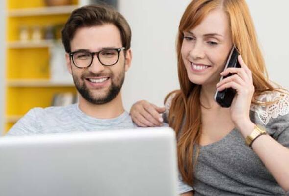 Two people sitting together using a laptop. One of them talking on phone.