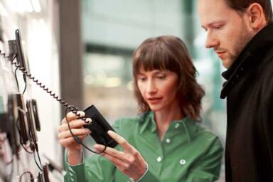 woman in green shirt showing man in black shirt a phone device attached to the wall