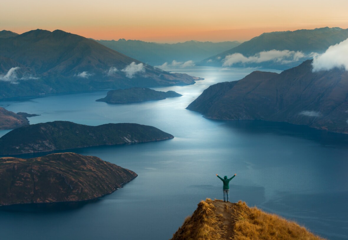 A view from above - a man with uplifted arms, standing on a peak, overlooking a lake.