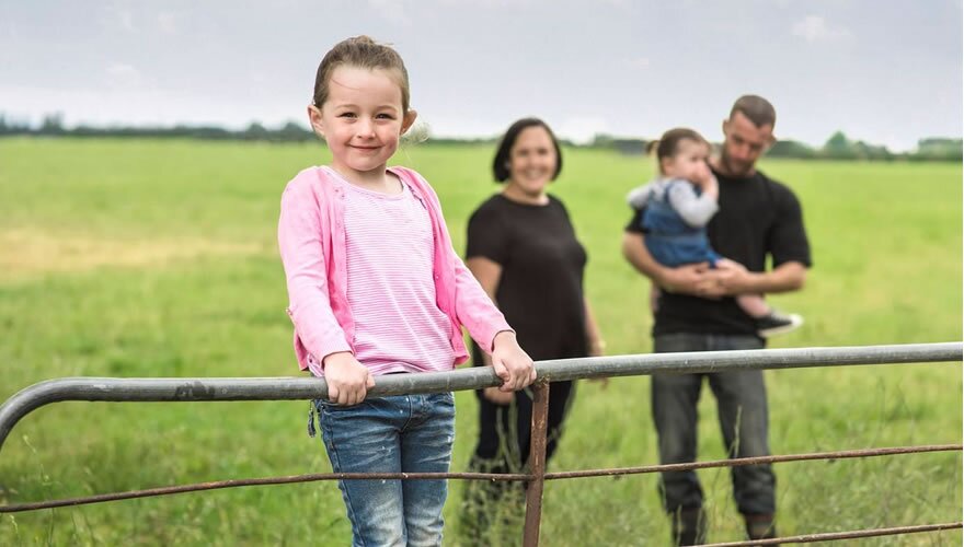Young girl in pink top and jeans standing on a farm gate with family behind her smiling.