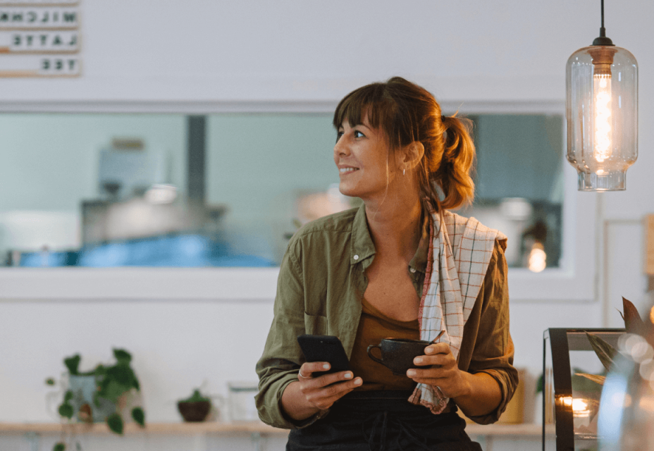 A smiling lady in a cafe, with a phone in one hand, and a cup of coffee in the other.